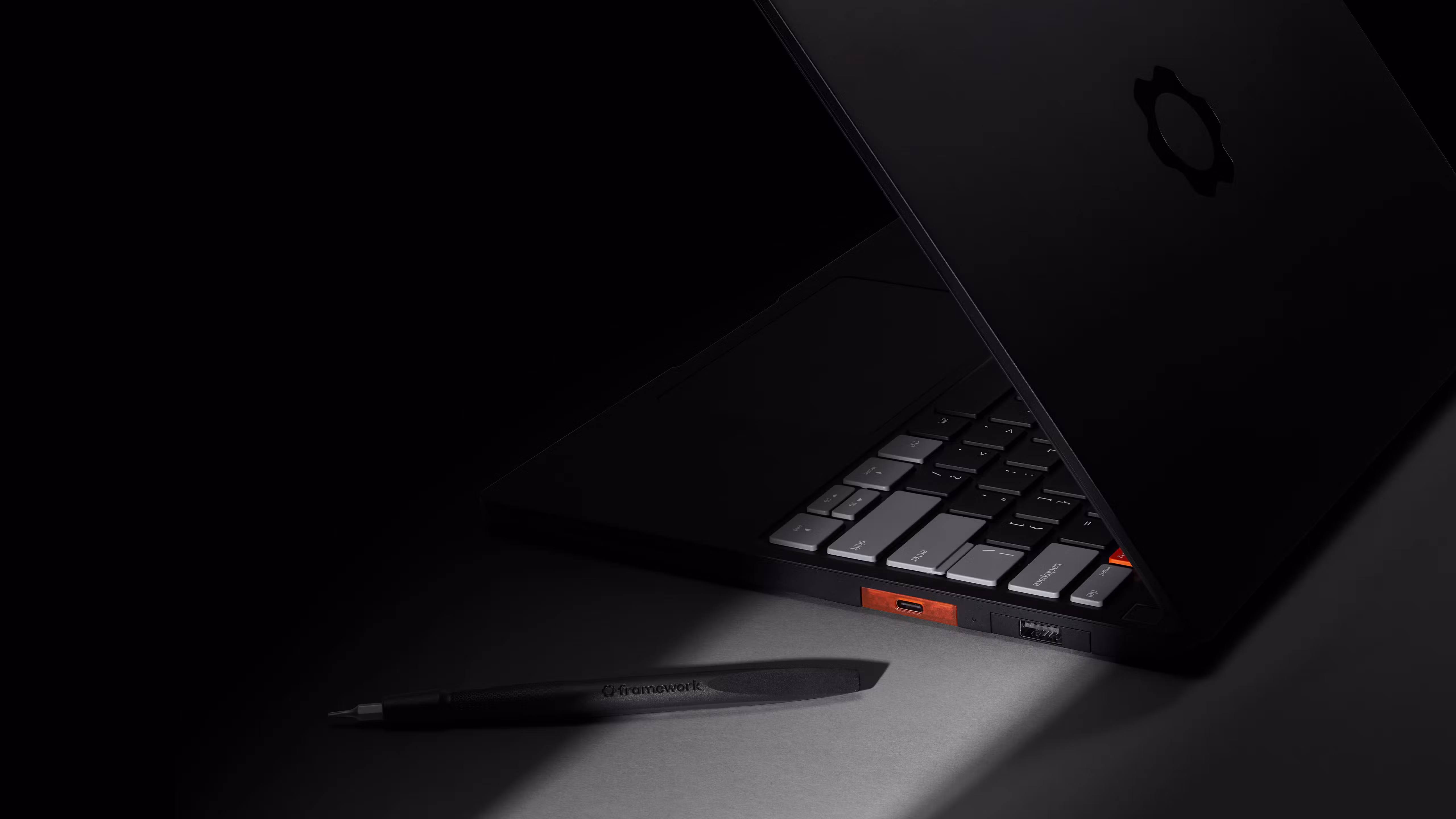 A high-angle, low-light shot of a sleek Framework Laptop 13 Pro partially open on a dark surface. A Framework screwdriver is placed closeby.