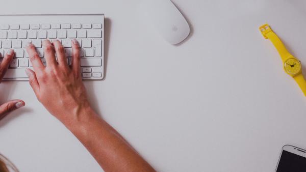 Manos escribiendo en un teclado con un reloj de pulsera y un teléfono celular al costado