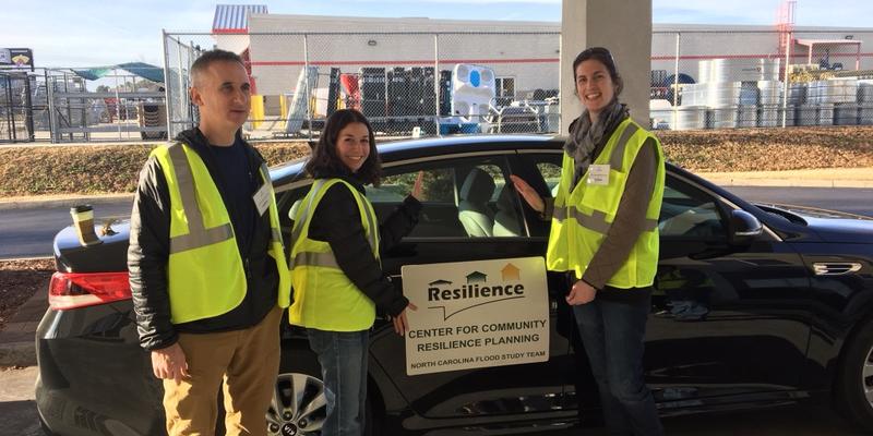 Three people in high-vis vests stand near a car that has a sign on the door: Center for Community Resilience Planning.